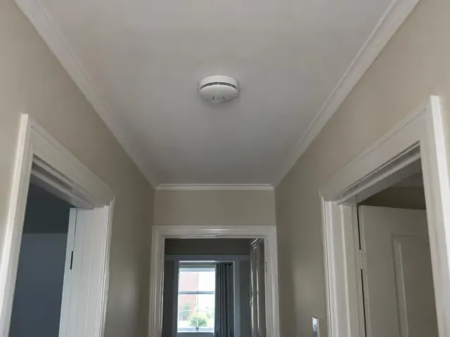 Ceiling-mounted smoke alarm in a painted hallway with door frames and a window at the end in East Maitland